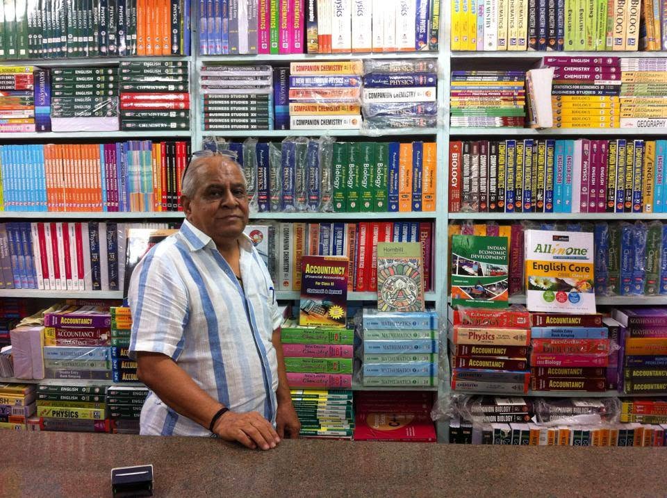 A bookseller standing behind the counter at Maneesh Book Shop, with shelves of school books behind him